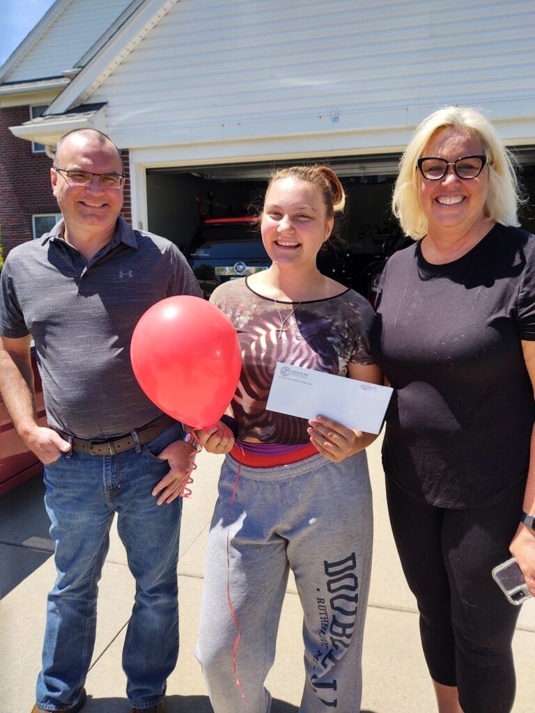 A girl holding a red balloon and a check stands with her parents.
