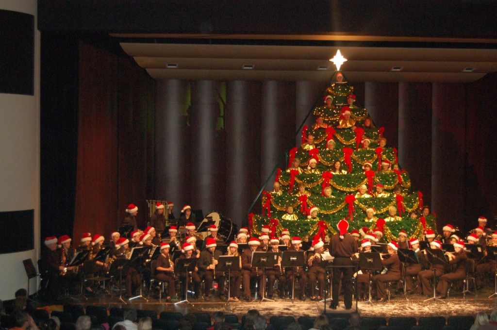 A large Christmas tree is surrounded by a band wearing Santa hats.
