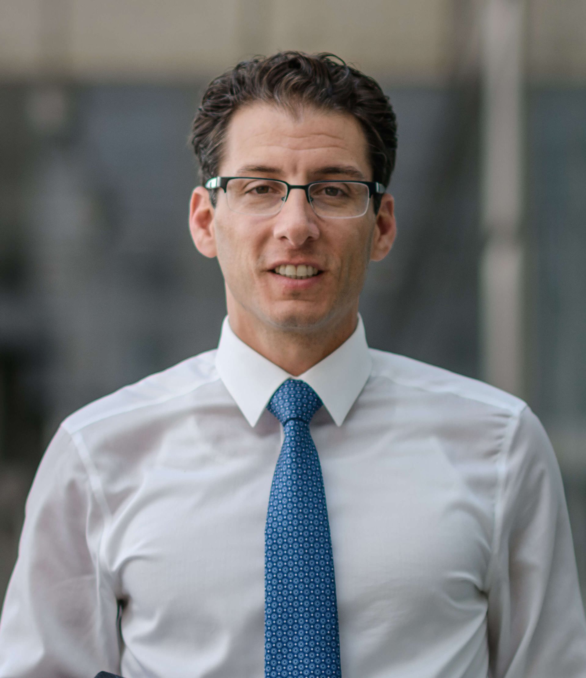 A man wearing glasses and a blue tie is smiling for the camera.