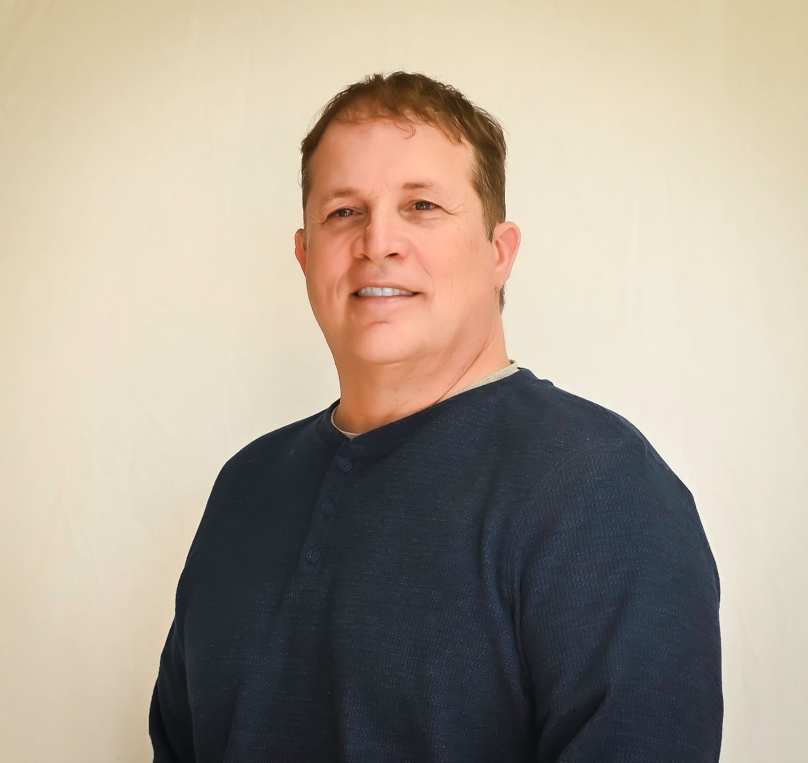 Man with short brown hair smiles, wearing a navy blue sweater against a beige wall.