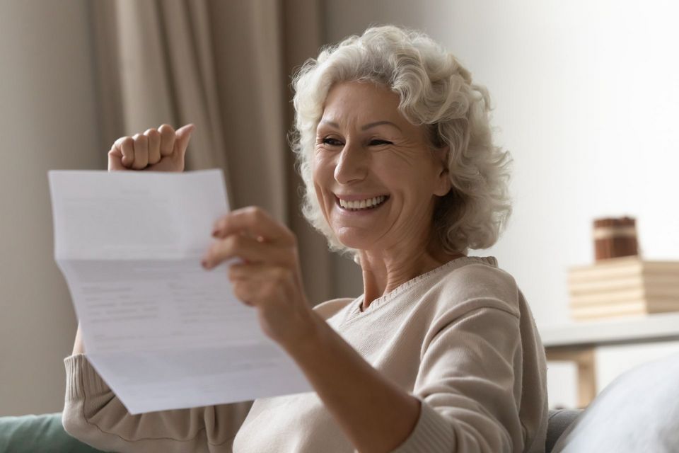 An elderly woman is smiling while holding a piece of paper.
