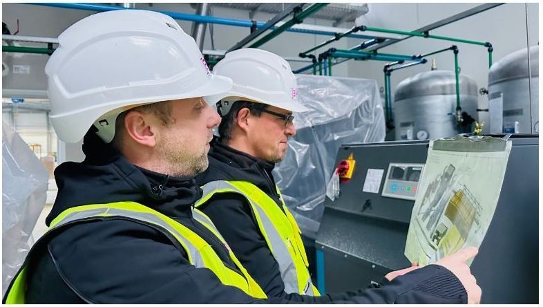 Zwei Männer mit Schutzhelmen und Sicherheitswesten schauen in einer Fabrik auf ein Klemmbrett.
