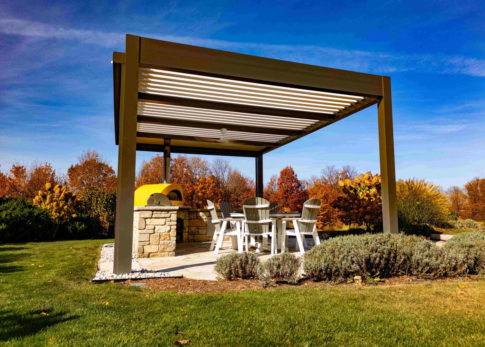 A dark brown louvered pergola over a stone outdoor pizza oven and patio dining set in a grassy yard under a blue sky.