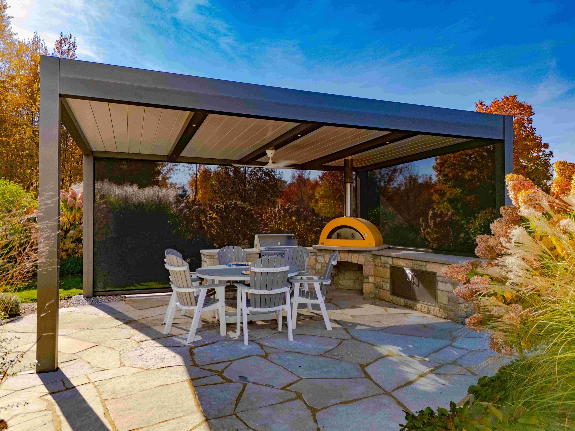 A patio features a modern pergola over a dining table, chairs, and a stone outdoor kitchen with a yellow pizza oven.