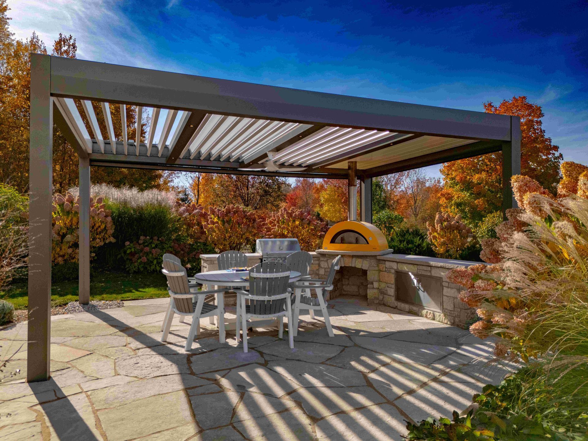 Outdoor patio with a table and chairs under a louvered pergola featuring a stone pizza oven in a fall landscape.
