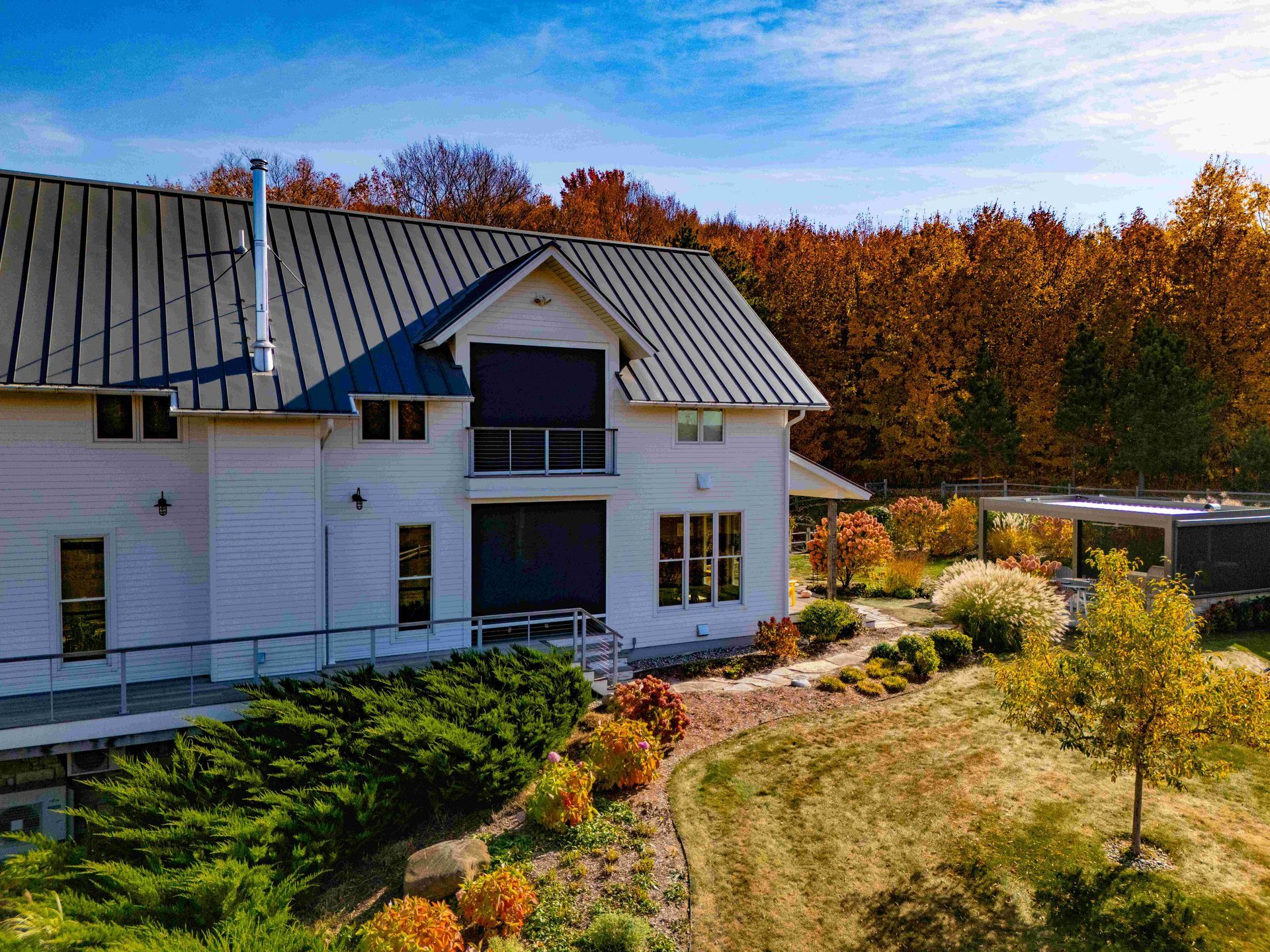 A white two-story house with a grey metal roof, surrounded by trees with autumn foliage and a grassy yard.
