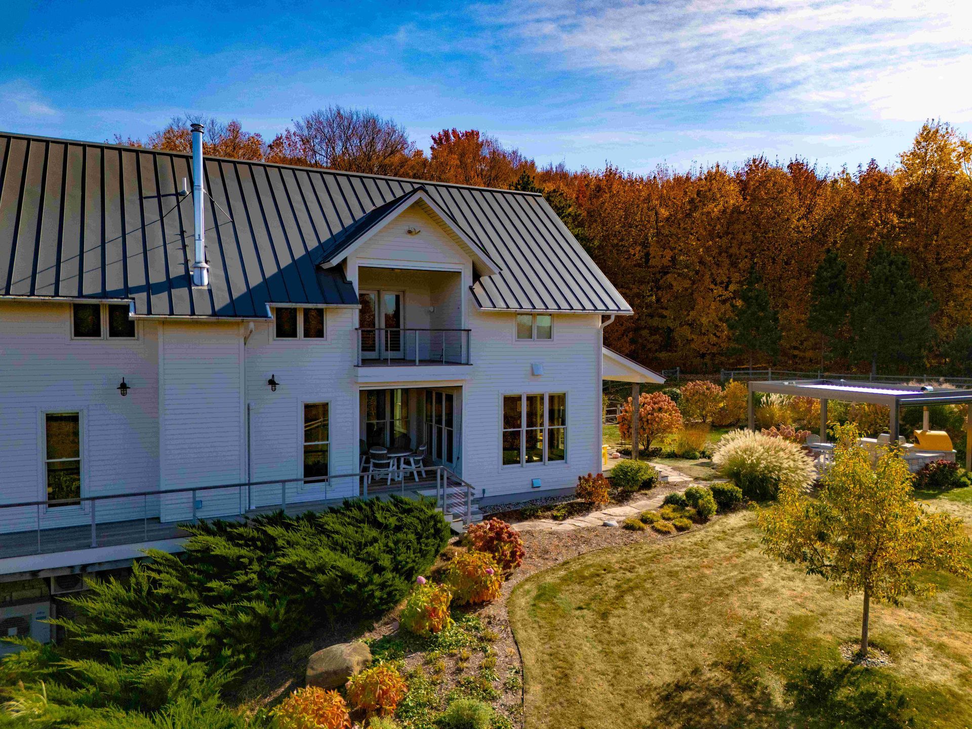 A white, two-story house with a metal roof surrounded by autumn foliage and a grassy yard under a bright blue sky.