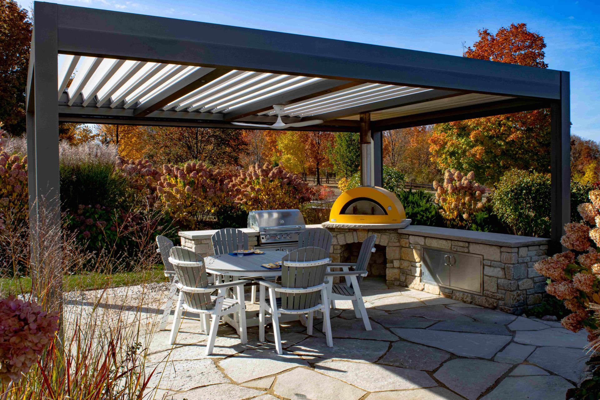 Outdoor patio with a pergola, dining set, stone countertop, and bright yellow pizza oven amidst autumn trees.