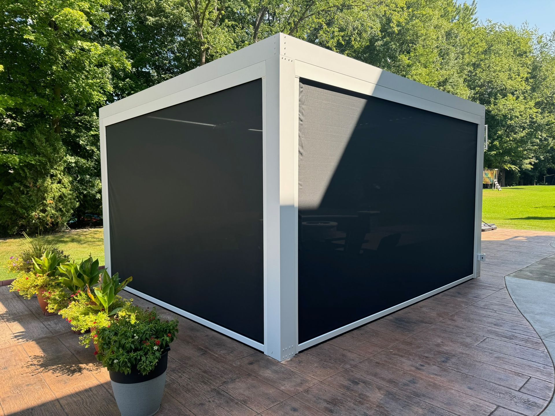 A white, modern pergola with closed black privacy screens, set on a stone patio with potted plants and trees in the back.