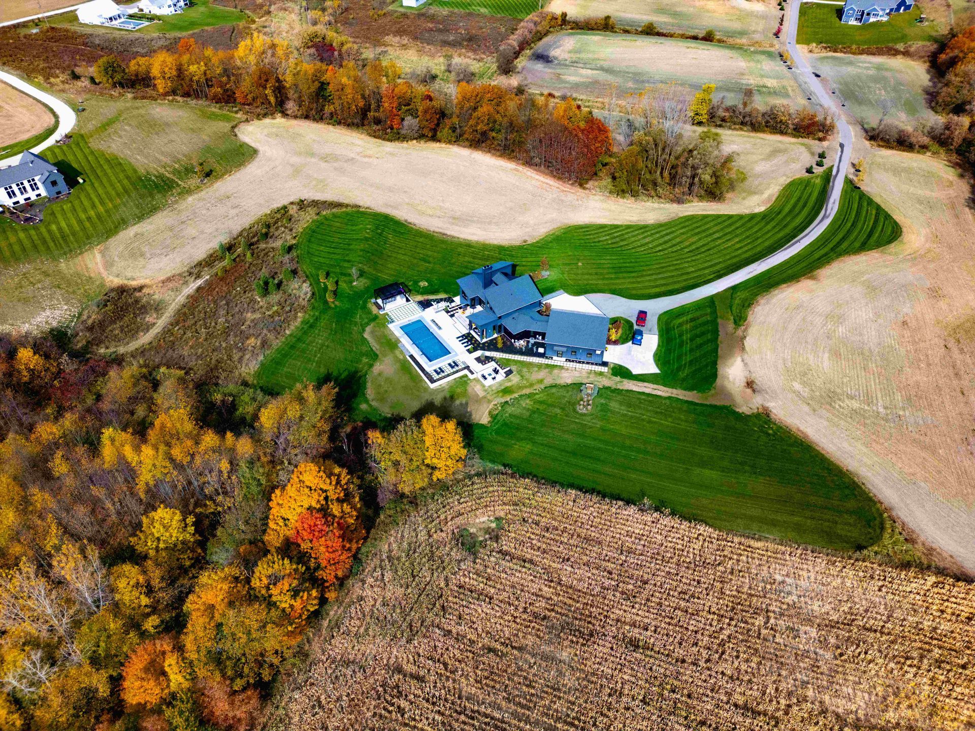 An aerial view of a blue house with a swimming pool, surrounded by fields and autumn trees in a rural landscape.
