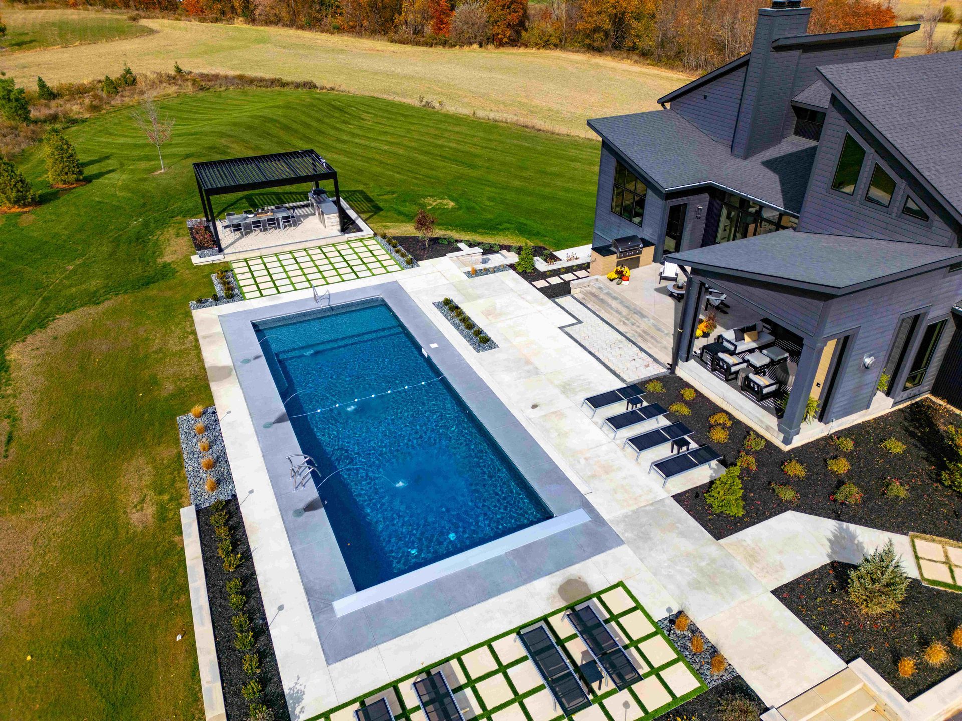 An aerial view of a modern house with a rectangular swimming pool, a patio, and a separate pergola in a grassy yard.