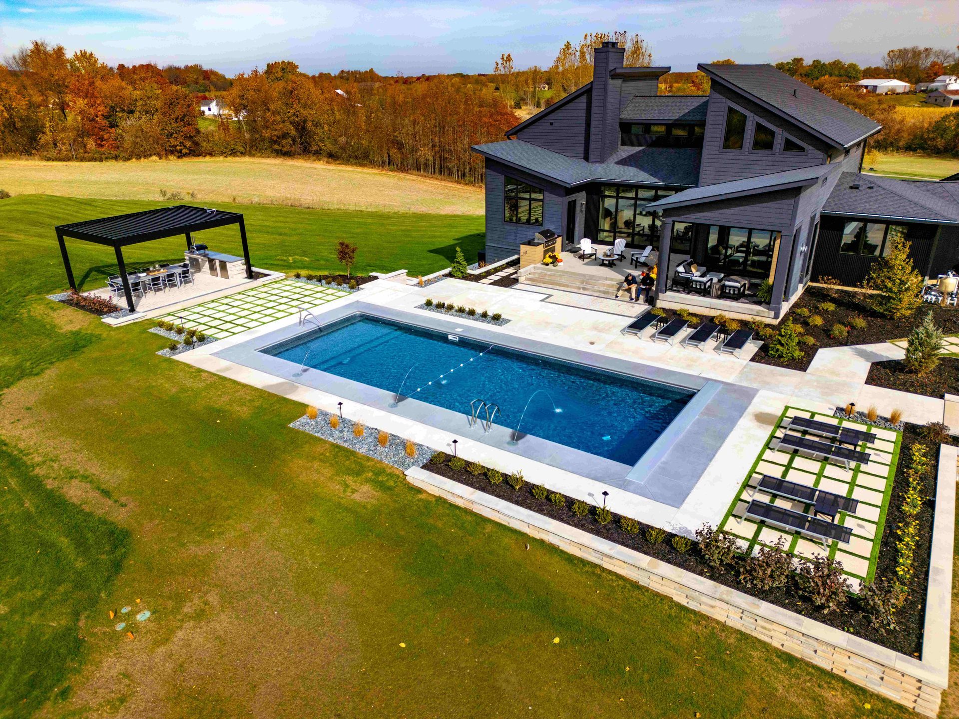 Aerial view of a modern dark-sided home with a rectangular pool, poolside loungers, and a covered patio in a grassy yard.