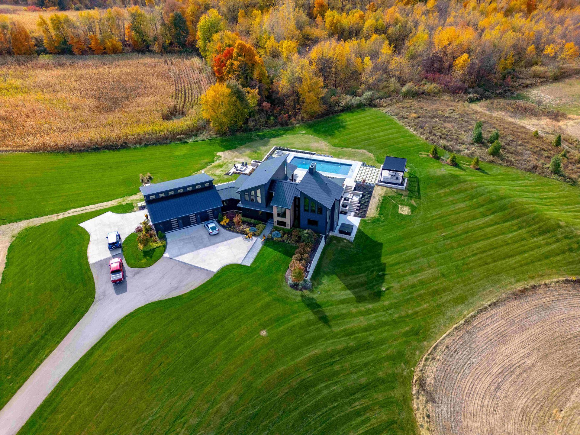 Aerial view of a modern dark-roofed home with a swimming pool, situated on a large grassy lot bordered by autumn trees.
