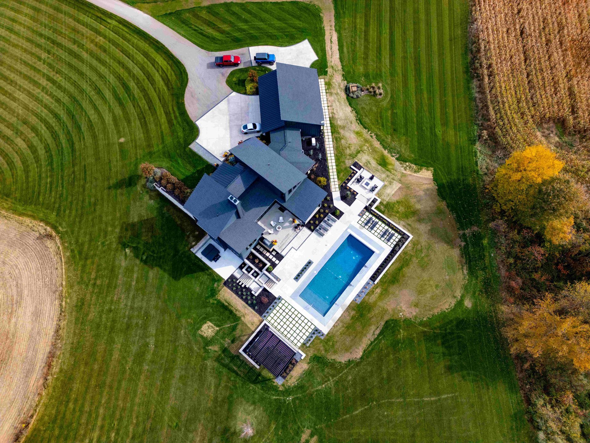 Aerial view of a house with a dark roof, a pool, and a patio, surrounded by a large lawn and a field of crops.