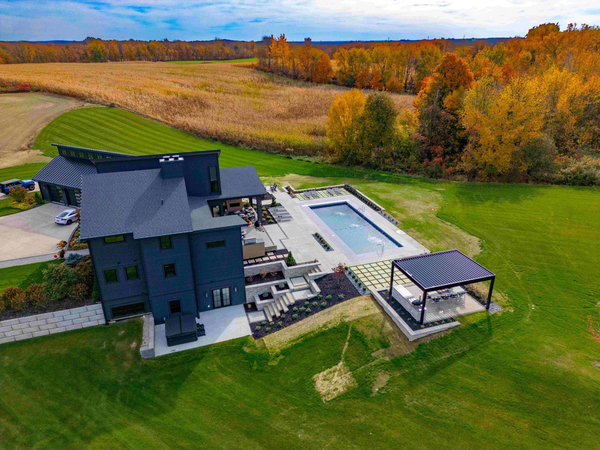 Aerial view of a modern dark-sided home with a swimming pool, patio, and pergola, set on a rural lawn near autumn woods.