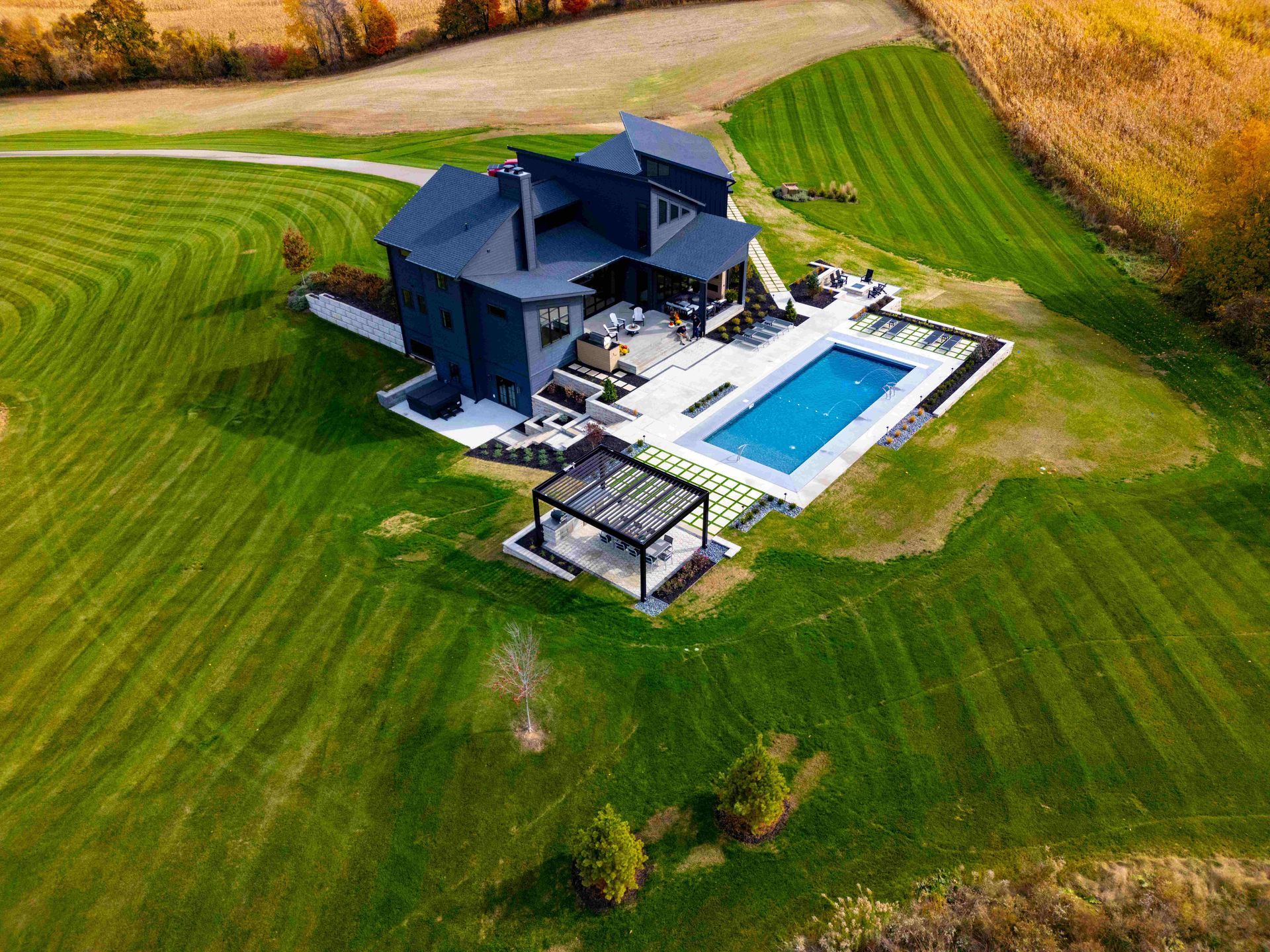 Aerial view of a dark blue multi-story modern home with a pool, patio, and pergola, set on a sprawling green lawn.