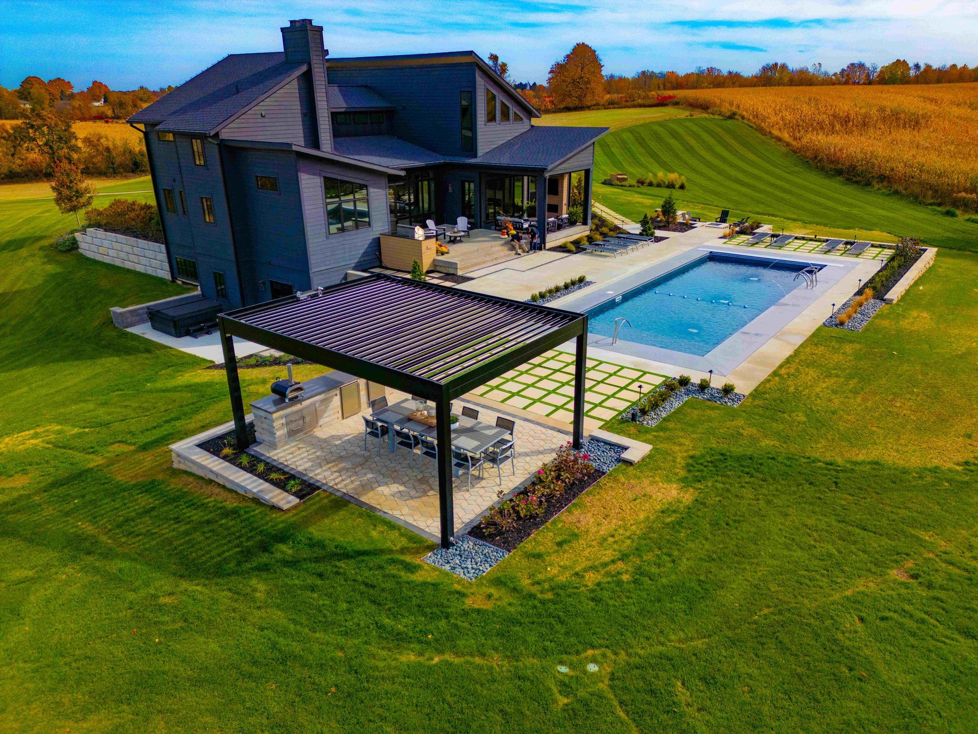 Aerial view of a dark gray house with a backyard pool, pergola-covered outdoor dining area, and surrounding lawn.