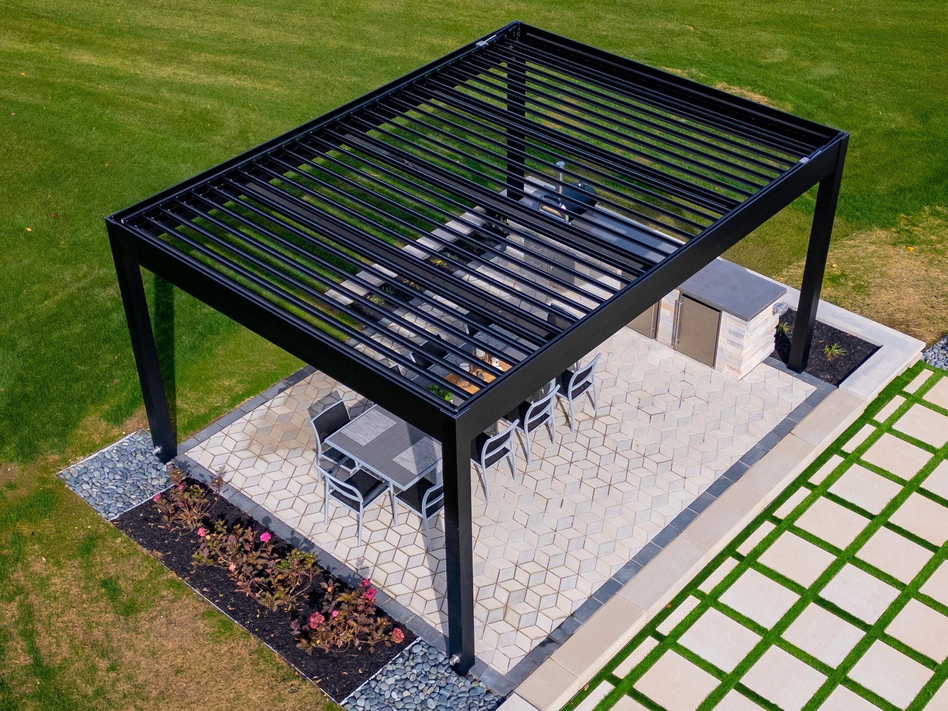 Aerial view of a black louvered pergola over a patio with dining furniture and a grill on a gravel base in a lawn.
