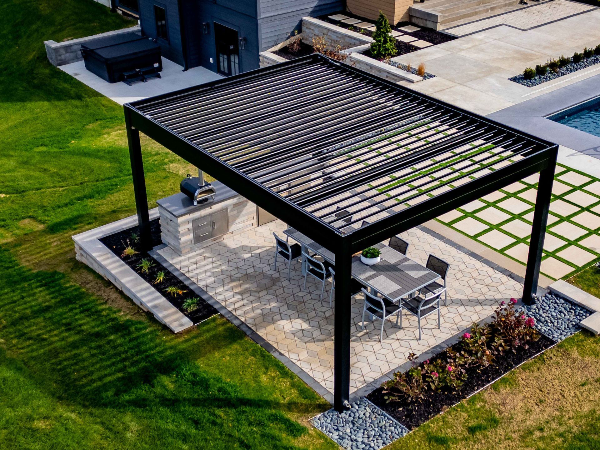 High-angle shot of a black louvered pergola over a stone patio with a dining table, chairs, and a grill in a backyard.