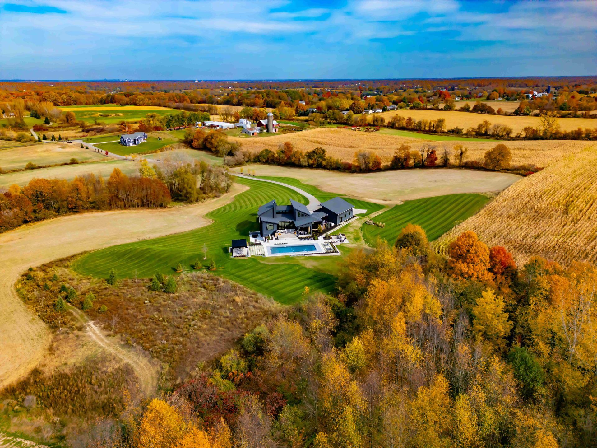 An aerial view of a dark-roofed modern house with a pool, surrounded by green lawns and autumn fields under a blue sky.