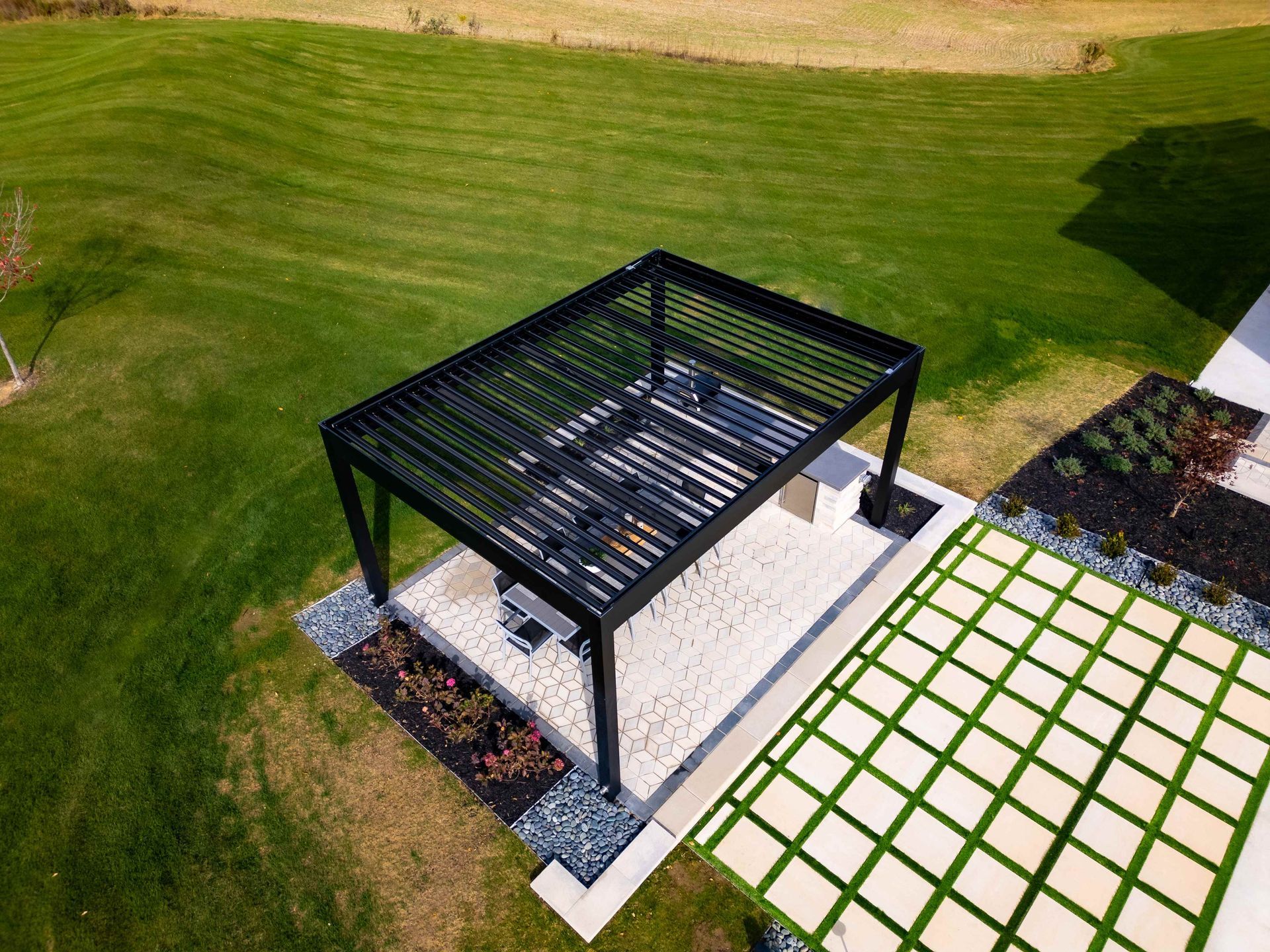 A black louvered pergola sits on a white gravel patio next to a square grid stone walkway in a lush green backyard.