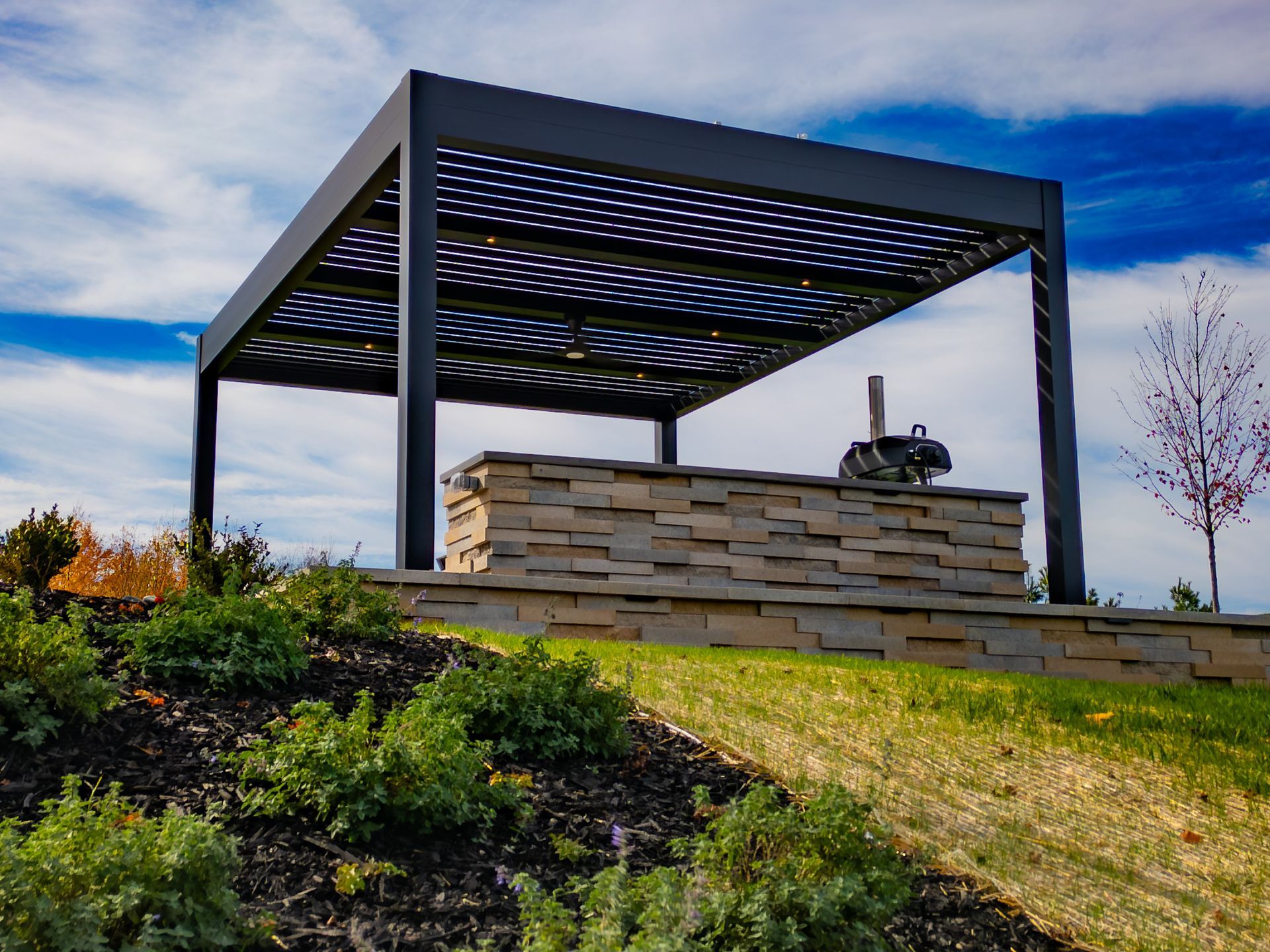A stone outdoor kitchen with a grill under a modern black pergola in a landscaped yard against a cloudy blue sky.