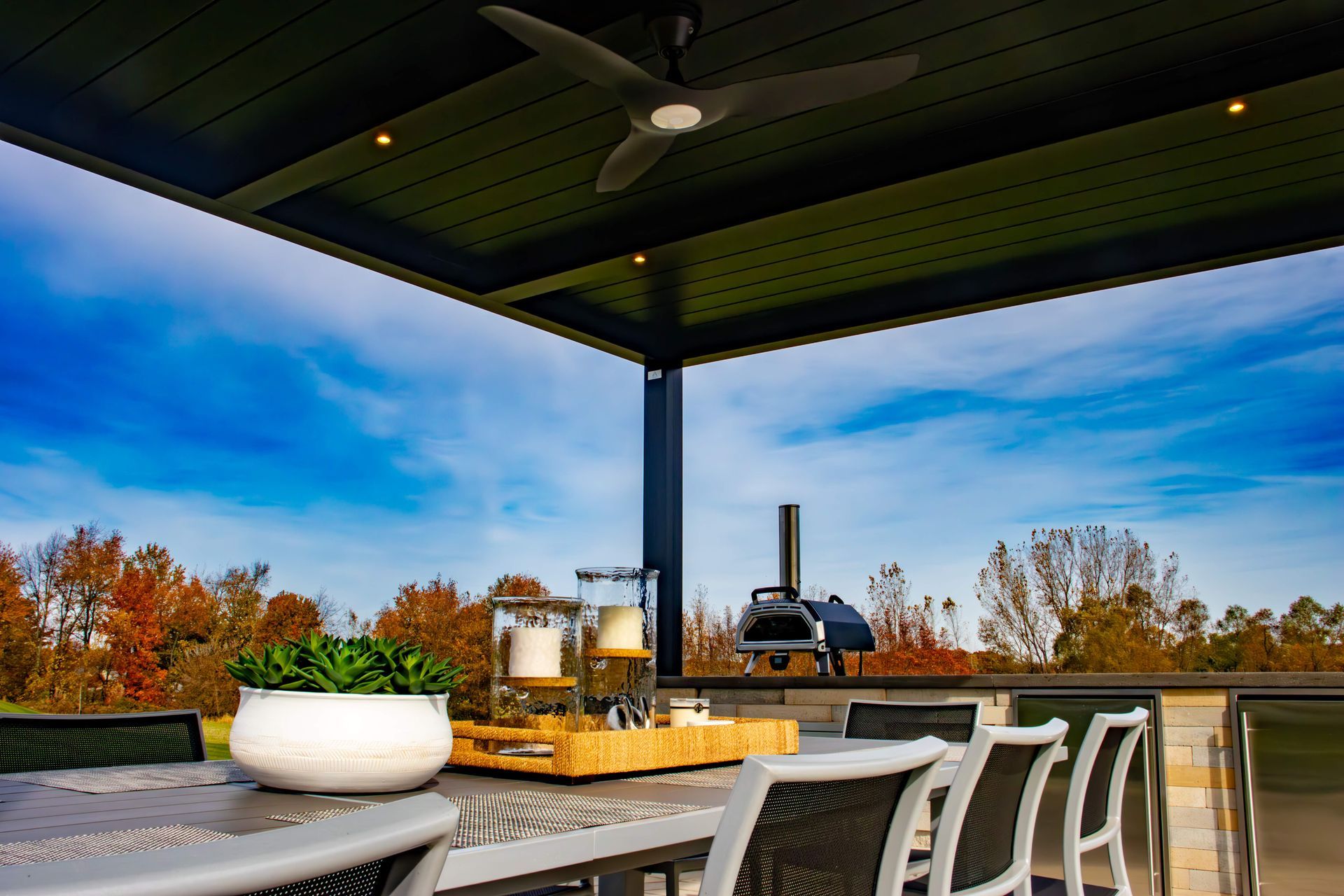 A covered outdoor dining patio featuring a dining table with a plant, candles, and a pizza oven under a dark ceiling fan.