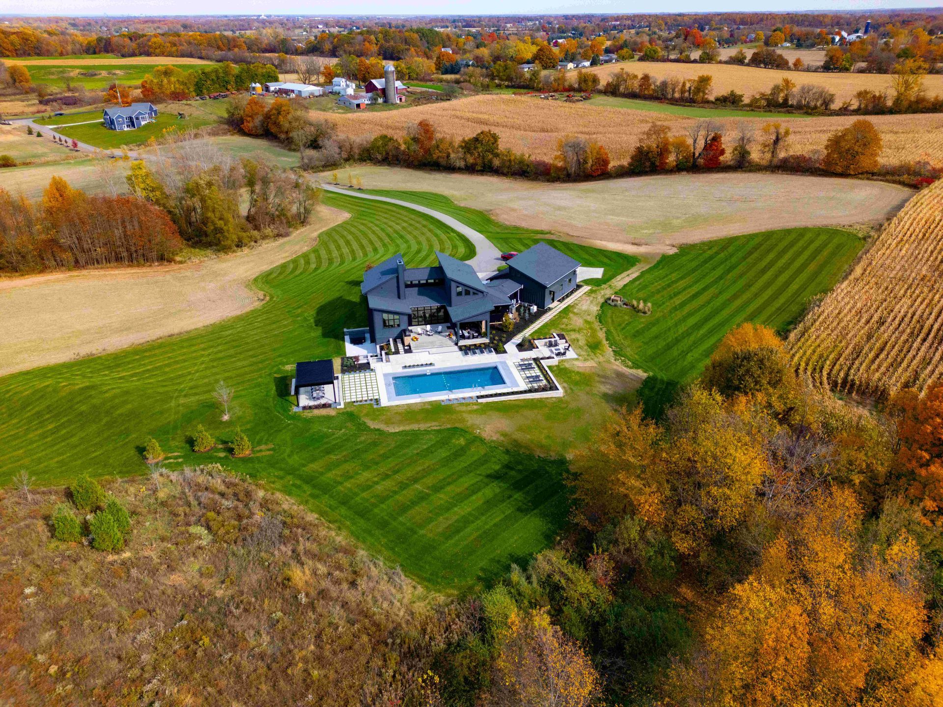 Aerial view of a modern dark-roofed house with a swimming pool, surrounded by expansive green lawns and autumn fields.