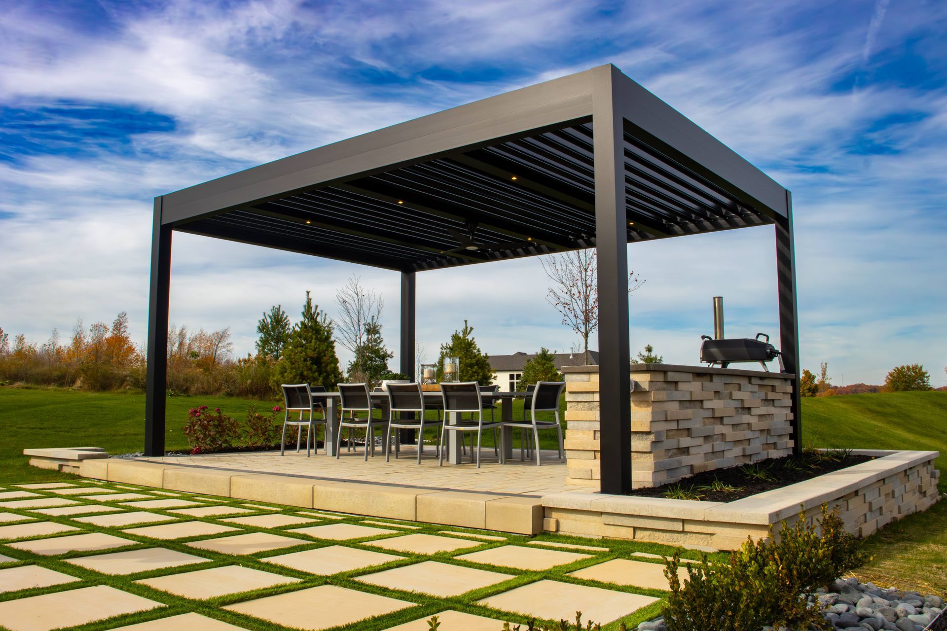 A modern black pergola covers an outdoor dining set and a stone kitchen island on a patio with a grassy landscape background.