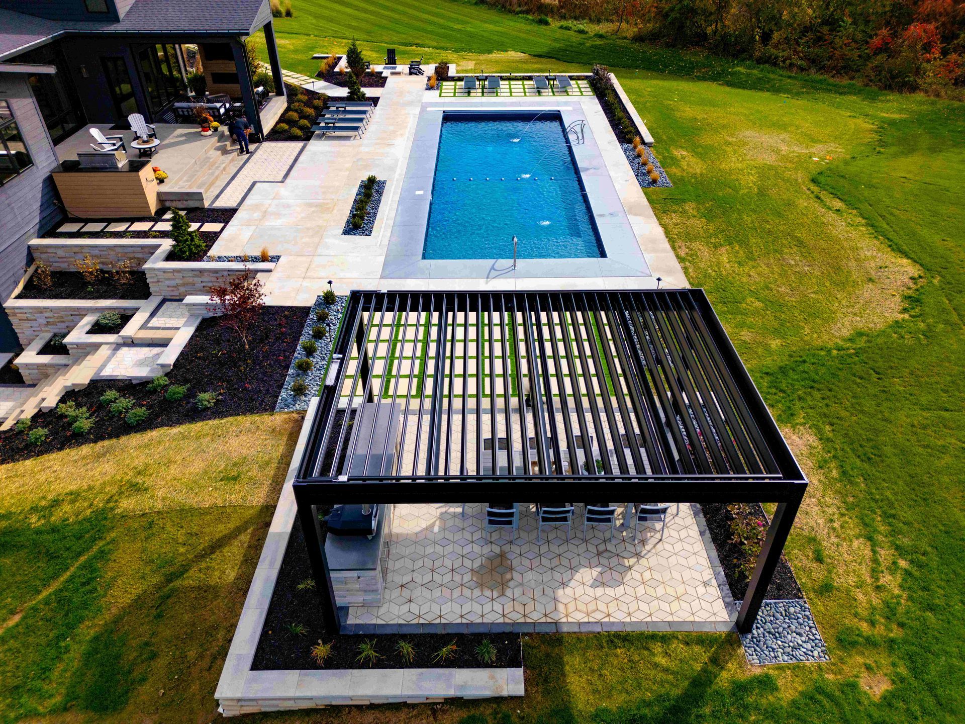 Aerial view of a backyard with a rectangular pool, a louvered pergola covering a patio, and tiered stone landscaping.