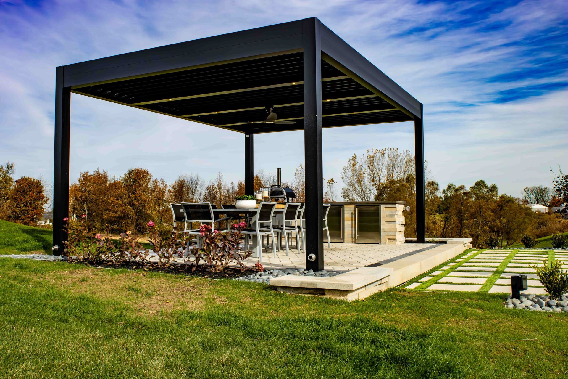 A modern black pergola covers a patio dining set and outdoor kitchen on a stone base, set in a sunny, grassy yard.