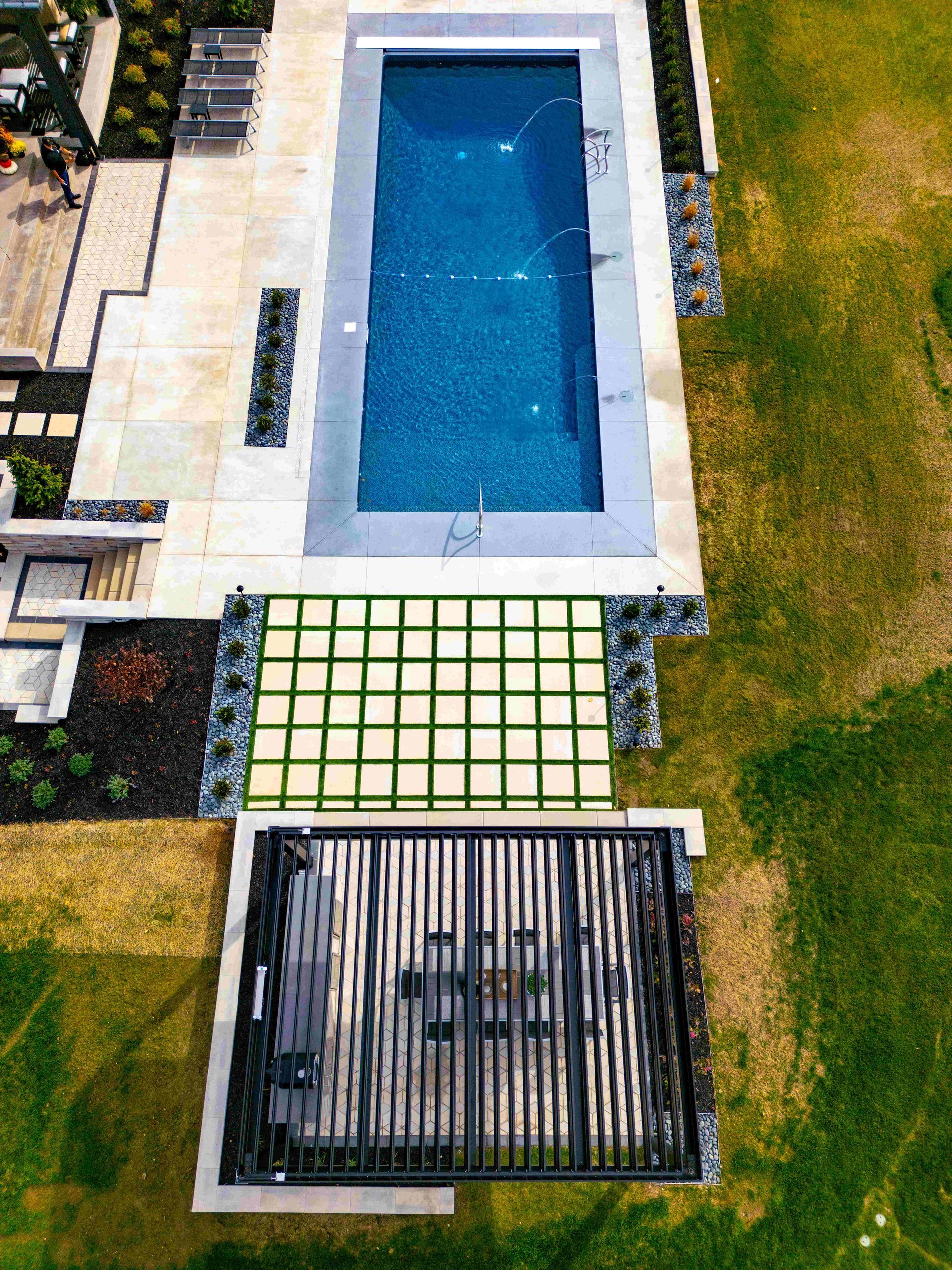 An aerial view of a backyard featuring a rectangular blue pool, a grid-patterned patio, and a shaded outdoor seating area.