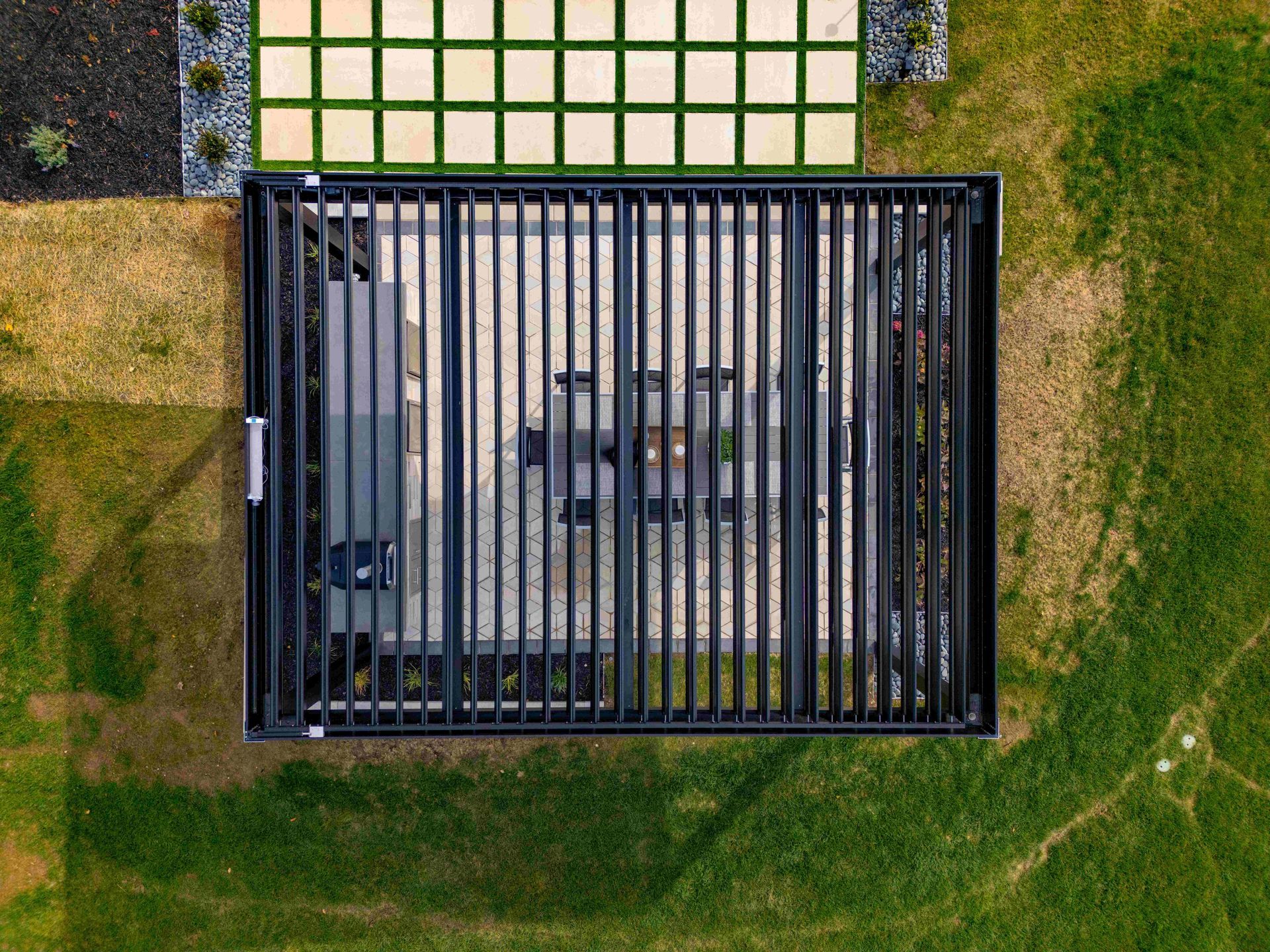 Top-down aerial view of a black louvered pergola over a patio, next to a grid-patterned stone path and grassy yard.