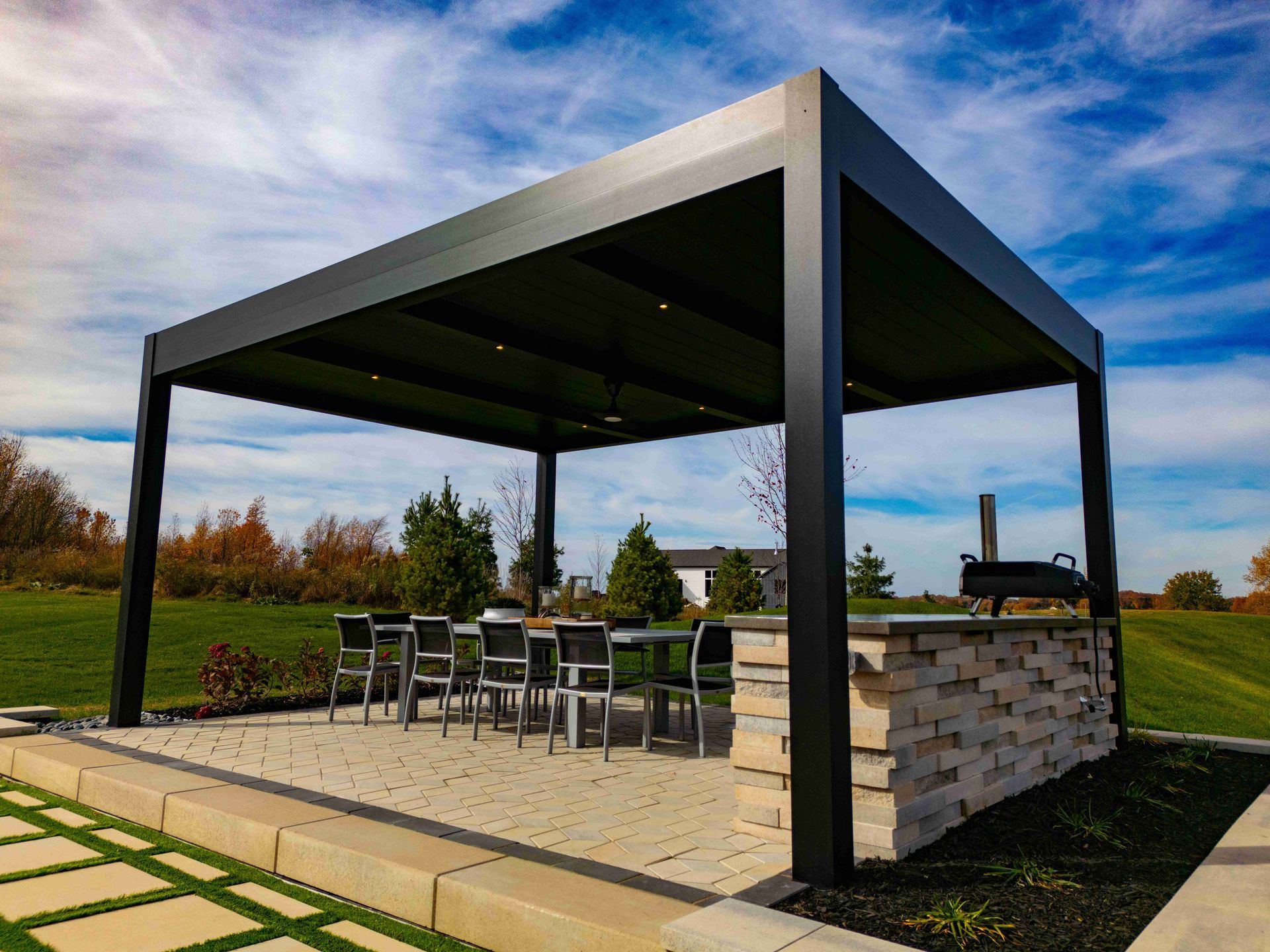 A modern black pergola covers an outdoor dining set and stone-built kitchen area on a paved patio in a grassy yard.
