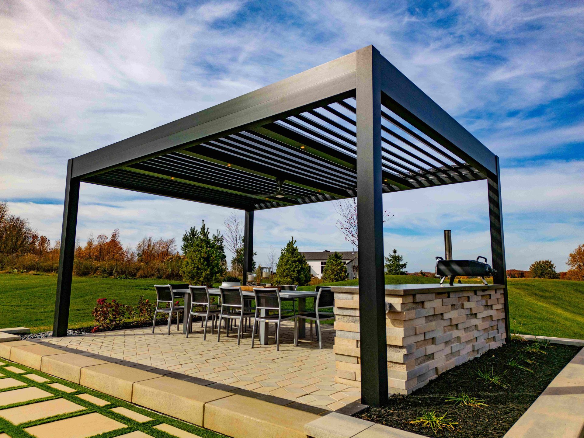 A modern black louvered pergola over a stone patio with outdoor dining furniture and a stone-built grill station.