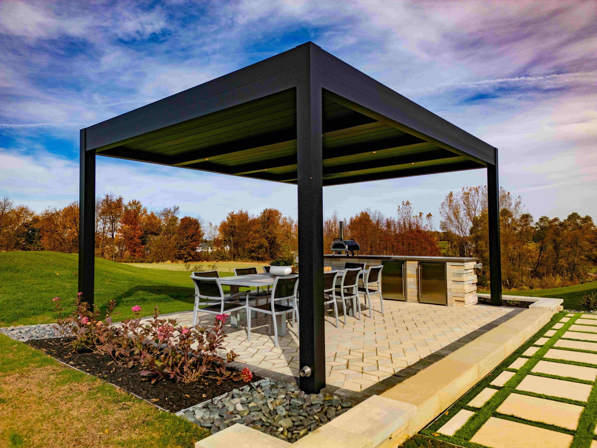 A modern black pergola covers an outdoor dining set and stone kitchen area on a patio set against a rural landscape.