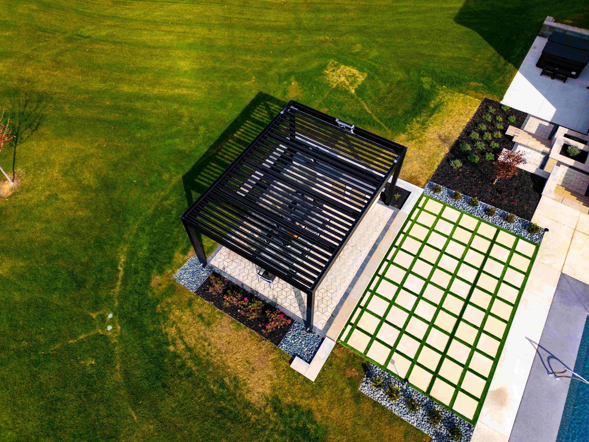 An aerial view of a black pergola over a stone patio, adjacent to a grid-patterned paver walkway on a grassy lawn.