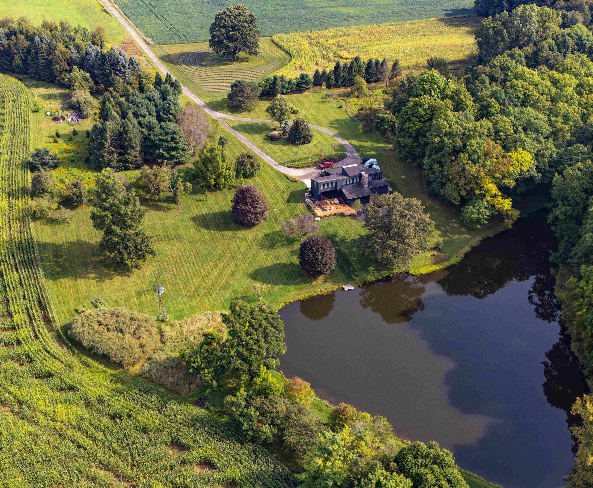 Aerial view of a dark-roofed house nestled in a lush, grassy yard with a private pond and surrounding trees.