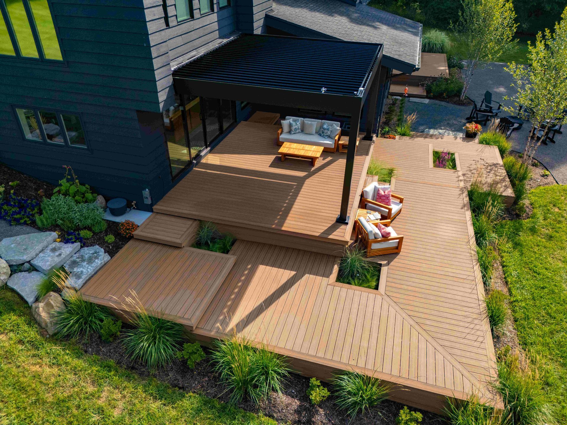 Aerial view of a tiered wooden deck with a black pergola, outdoor furniture, and surrounding greenery next to a dark home.