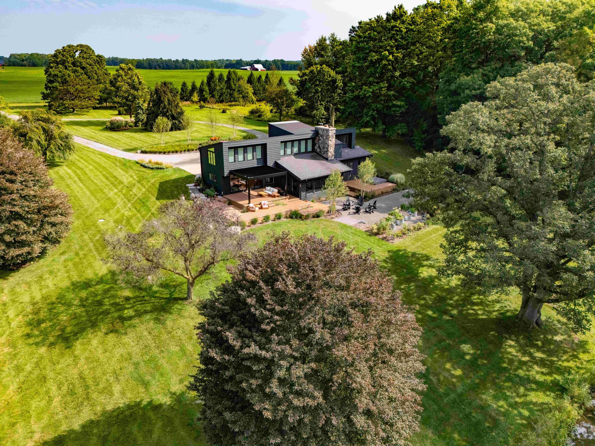 An aerial view of a dark, modern house nestled in a lush, green, wooded landscape under a clear blue sky.