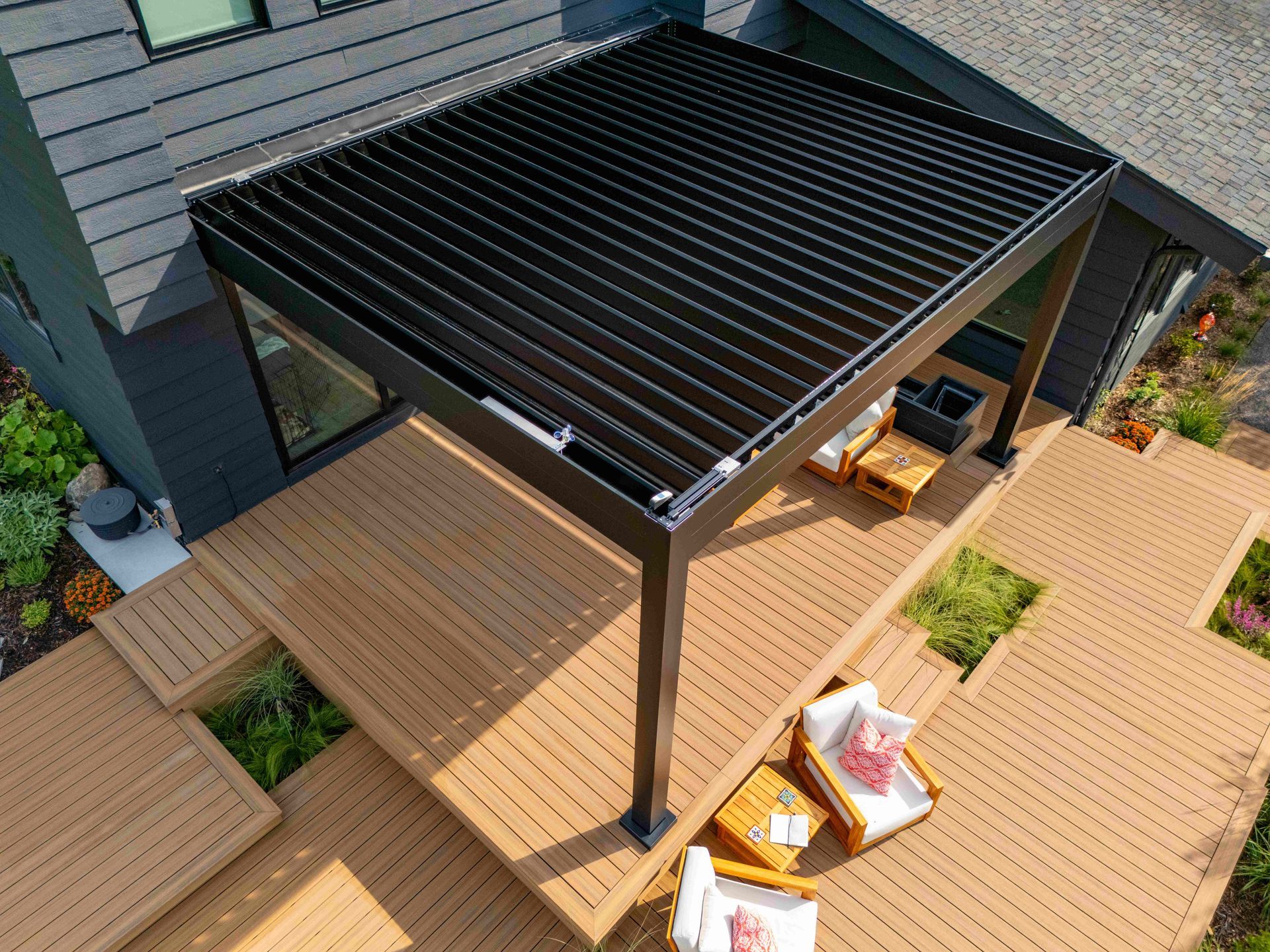 A black louvered pergola structure installed over a wooden deck with outdoor seating, viewed from a high angle.