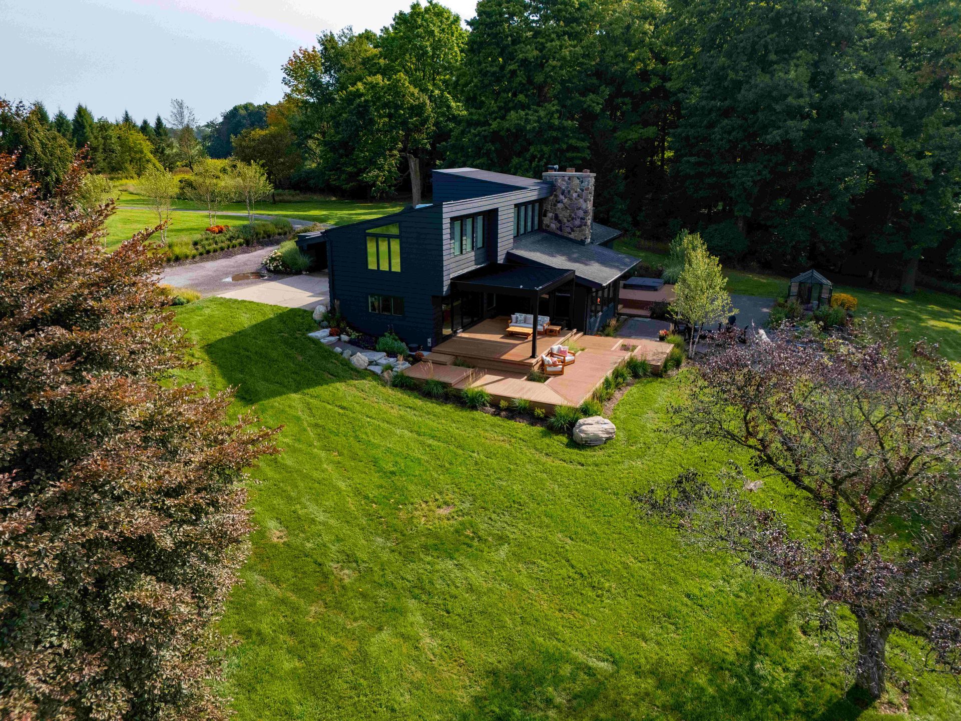 An aerial view of a dark, modern house with a stone chimney, surrounded by a lush green lawn and mature trees.