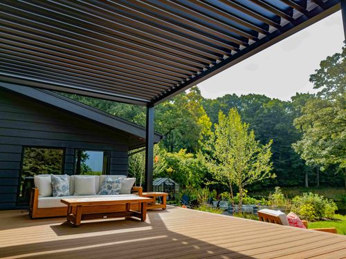 A deck with a louvered pergola, outdoor seating, and a coffee table, overlooking a lush backyard with mature trees.
