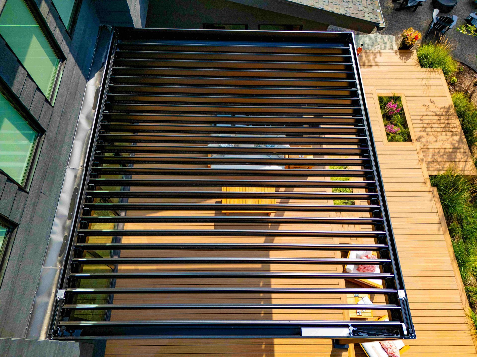 A high-angle view of a black louvered pergola roof installed over a wooden deck next to a modern building.