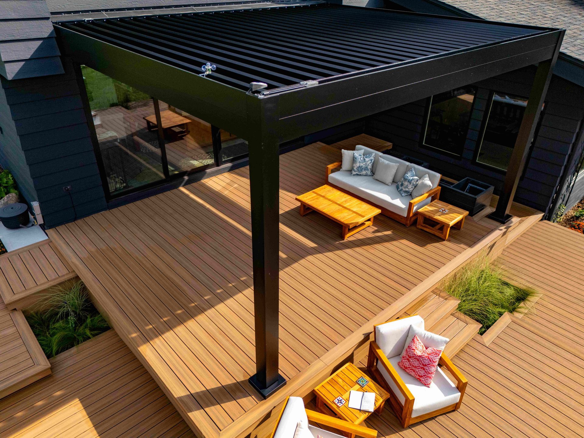 A black louvered pergola shades a wooden deck with a sofa, armchairs, and coffee tables, set against a modern house.