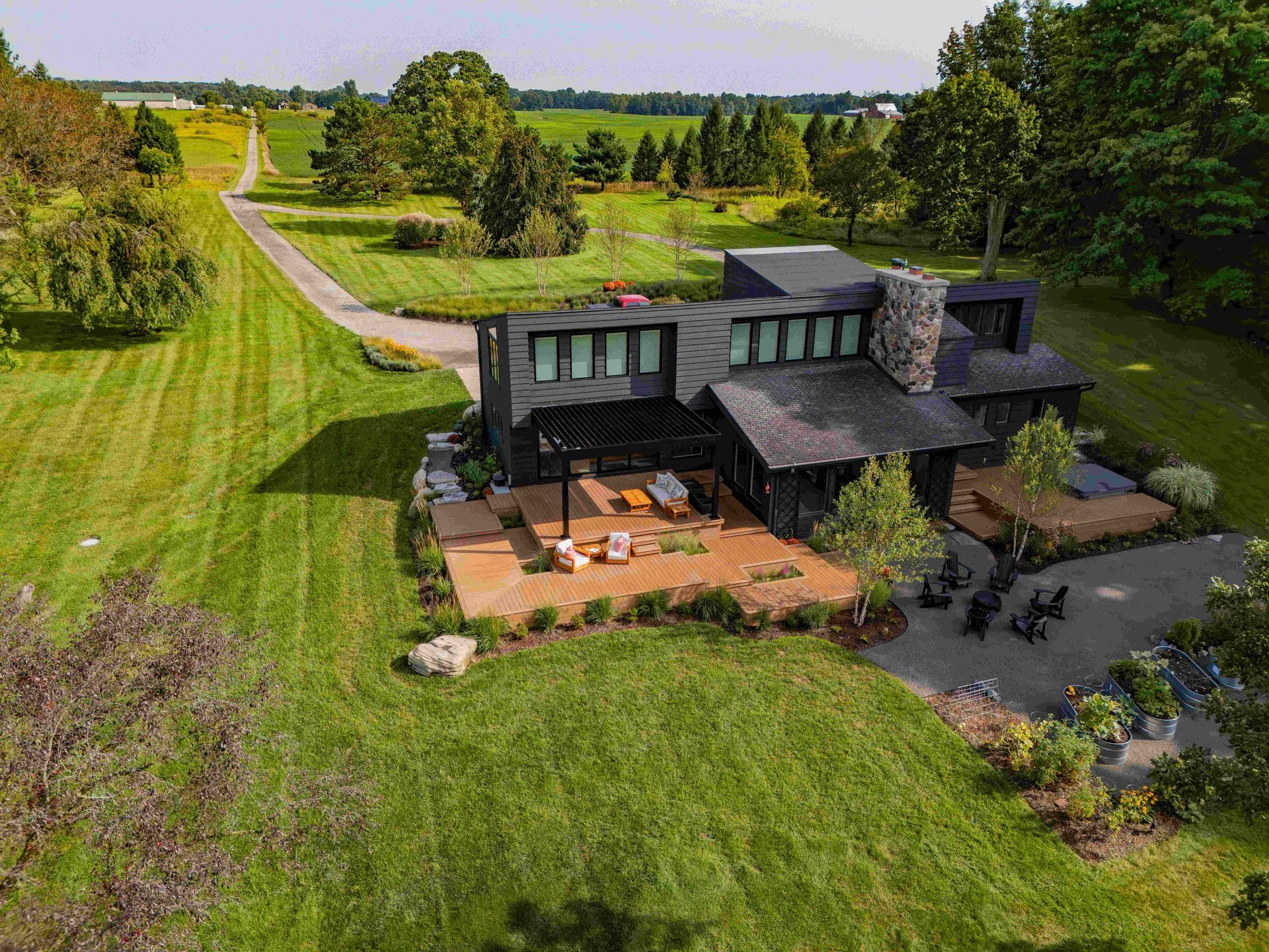 Aerial view of a modern black home with a stone chimney, outdoor patio, and deck surrounded by a large green field.