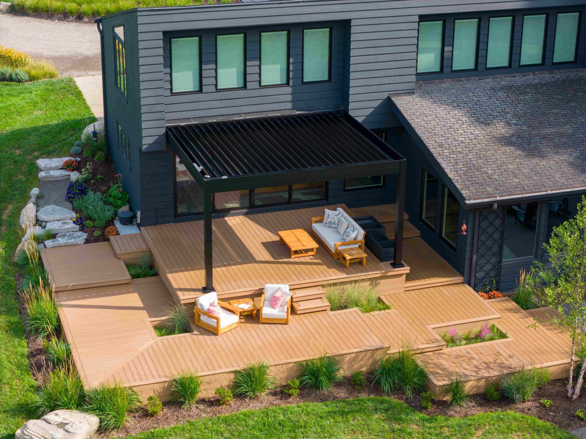Modern house with a tiered wooden deck, outdoor seating under a louvered pergola, and landscaped garden beds.