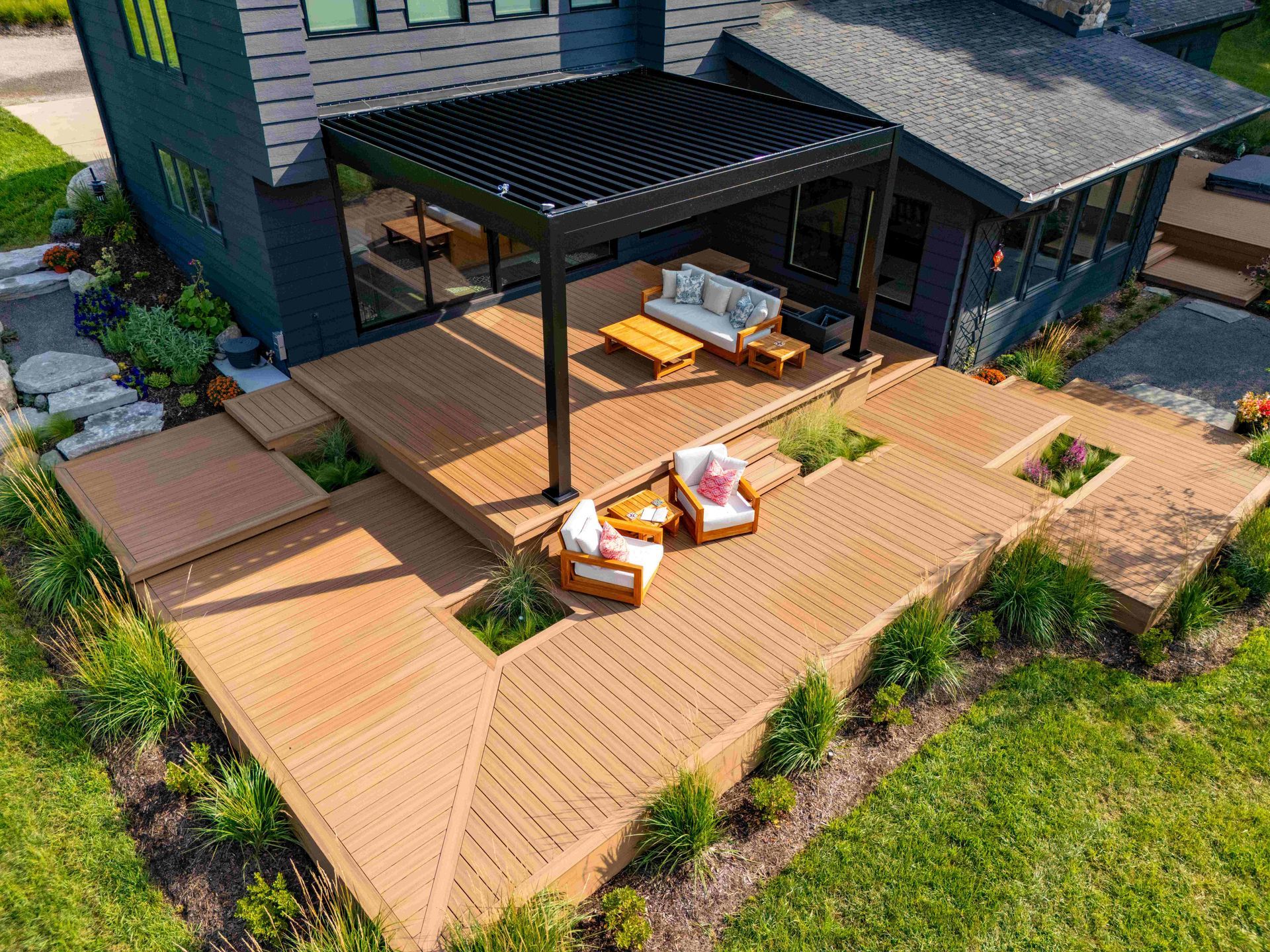 An aerial view of a multi-level wooden deck attached to a dark house, featuring a louvered pergola, sofas, and armchairs.