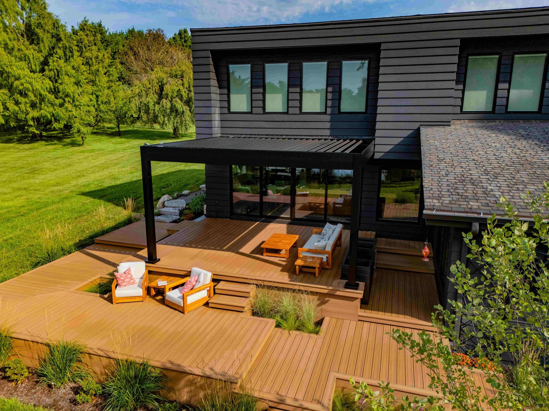 A modern home with a dark exterior, featuring a wooden deck, a pergola, and outdoor patio furniture set in a grassy yard.