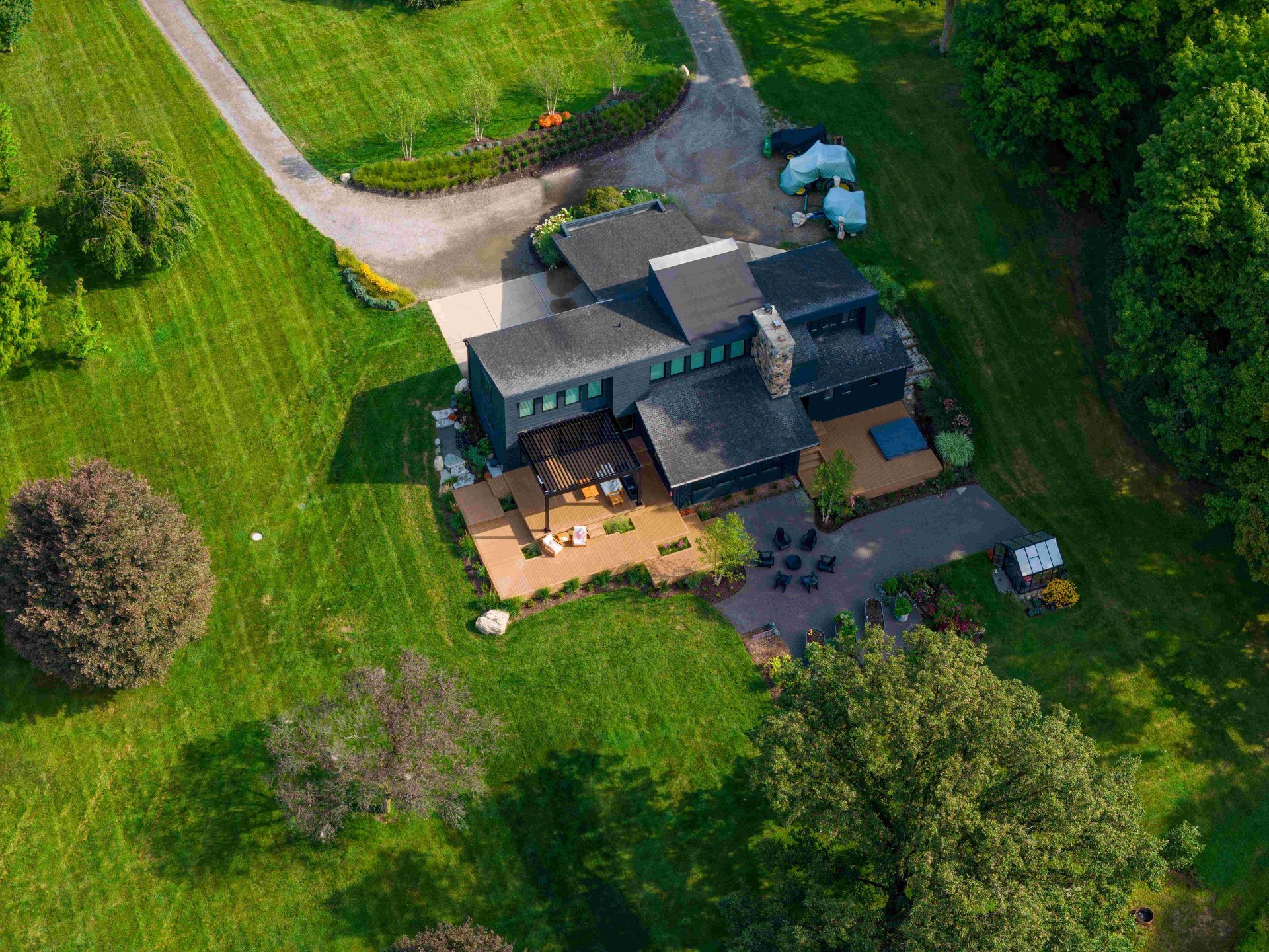 An aerial view of a dark-roofed home on a large, green grassy lot with a patio, driveway, and surrounding forest.